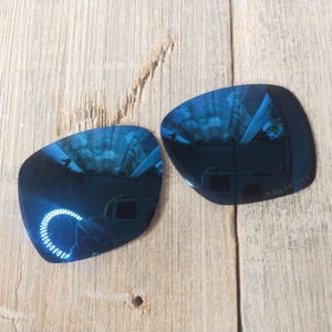 May include: Two blue mirrored replacement lenses for sunglasses. The lenses are curved and have a reflective blue coating. The text "POLARIZED" is printed on the bottom right corner of one lens.