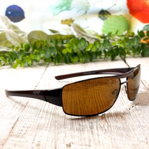 May include: Dark brown sunglasses featuring a rectangular frame design with amber-tinted lenses. The temples display a small metallic logo detail on the side, and the bridge includes adjustable nose pads for a secure fit on the face.