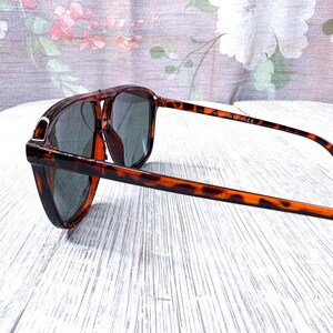 May include: Tortoiseshell sunglasses with dark lenses. The frames have a classic aviator shape with a double bridge and a brown and black pattern. The arms are slender and extend to the temples. The sunglasses are angled, showing the side view.