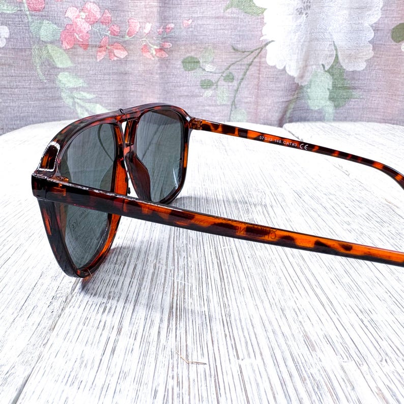 May include: Tortoiseshell sunglasses with dark lenses. The frames have a classic aviator shape with a double bridge and a brown and black pattern. The arms are slender and extend to the temples. The sunglasses are angled, showing the side view.