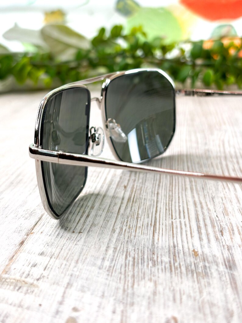 Pode incluir: &Oacute;culos de sol com arma&ccedil;&atilde;o prateada e lentes escuras. Os &oacute;culos de sol t&ecirc;m um formato aviador cl&aacute;ssico com ponte dupla e arma&ccedil;&atilde;o de metal fina. As lentes s&atilde;o de cor cinza escuro e a arma&ccedil;&atilde;o tem um acabamento brilhante.