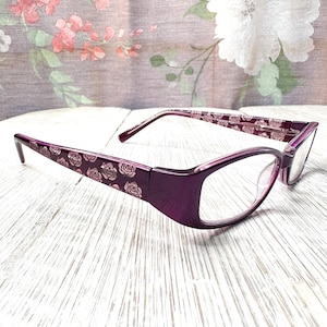 Puede incluir: Gafas moradas con montura rectangular. Las patillas presentan un estampado de rosas y la montura tiene un acabado brillante. Las lentes son transparentes y las gafas están sobre un fondo floral.