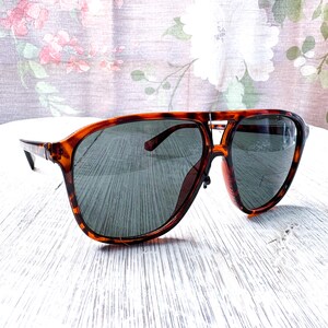 May include: Tortoiseshell sunglasses with dark lenses. The sunglasses have a classic aviator style with a double bridge and a thick frame. The lenses are a dark gray color, and the frame has a brown and amber pattern.