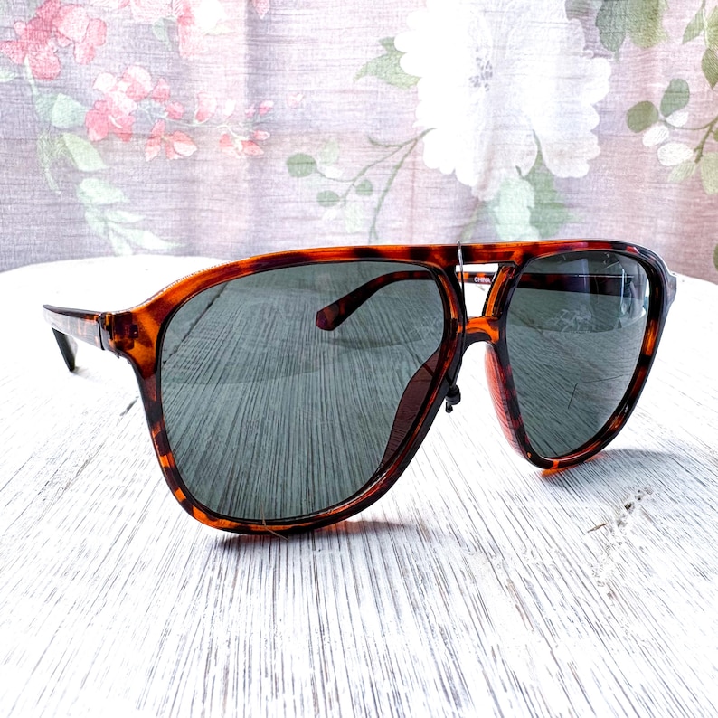 May include: Tortoiseshell sunglasses with dark lenses. The sunglasses have a classic aviator style with a double bridge and a thick frame. The lenses are a dark gray color, and the frame has a brown and amber pattern.