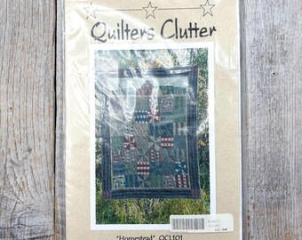 Könnte beinhalten: Eine Packung für ein Quiltprojekt, mit einem kleinen Quilt mit einem Patchwork-Design in Grün-, Rot- und Blautönen. Die Packung ist mit "Quilters Clutter" beschriftet und enthält den Text "Homestead" und "Designed for Diamond Textiles."