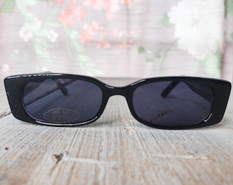 May include: Black rectangular sunglasses with dark lenses. The sunglasses have a thick frame and are resting on a wooden surface. The lenses have a UV protection sticker.