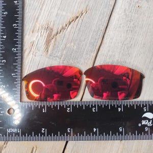 Puede incluir: Dos lentes de repuesto de color rojo espejado para gafas de sol, medidas con una regla.