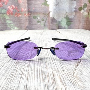 May include: Rimless sunglasses with purple lenses and black temples. The lenses have a rounded oval shape and the frame is a dark metal. The text "MAXIMUM PROTECTION UV400" is printed on the right lens.