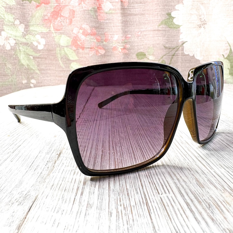 以下が含まれることがあります： ダークブラウンのフレームと紫色のグラデーションレンズを備えた特大サングラス。 サングラスは正方形の形状と太いフレームです。 レンズは上部で濃い色合いから下部で明るい色合いに移行します。