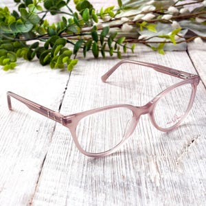Może przedstawiać: Jasnoróżowe okulary w kształcie kociego oka. Oprawki mają subtelną, przezroczystą jakość, a zauszniki mają dekoracyjny detal. Soczewki są przezroczyste, a okulary spoczywają na białej drewnianej powierzchni z zielenią w tle.