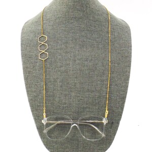 Op de afbeelding: Gouden ketting brillenhouder met drie zeshoekige bedels. De ketting hangt aan de nek van een mannequin en een heldere bril is aan de ketting bevestigd.