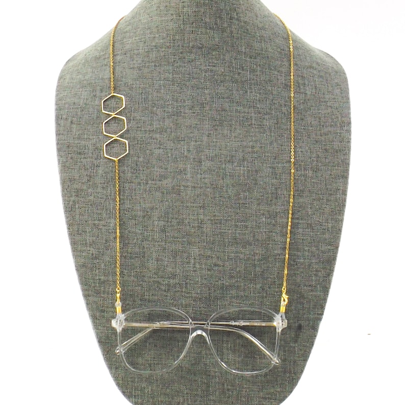 Op de afbeelding: Gouden ketting brillenhouder met drie zeshoekige bedels. De ketting hangt aan de nek van een mannequin en een heldere bril is aan de ketting bevestigd.
