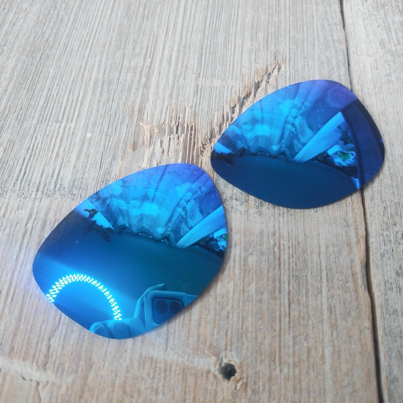May include: Two blue mirrored replacement lenses for sunglasses. The lenses are oval shaped and have a reflective blue coating.