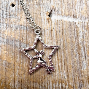 Puede incluir: Collar plateado con un colgante en forma de estrella. El colgante tiene un acabado texturizado y presenta una figura de hada dentro de la estrella. La cadena es plateada, sobre un fondo de madera rústica.