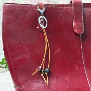 May include: A close-up of a red leather tote bag with a silver keychain and a decorative tassel. The tassel features small, colourful beads. The bag has a handle and a flap closure.