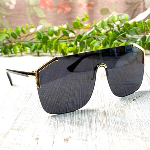 Può includere: Occhiali da sole neri con montatura in metallo color oro. Gli occhiali da sole hanno un design a scudo con lenti scure e un aspetto elegante e moderno. La montatura ha un accento dorato sulla parte superiore e sui lati. Le aste sono nere.