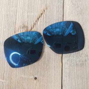 May include: Two blue polarised replacement lenses for sunglasses. The lenses are curved and have a reflective surface.