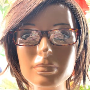 Puede incluir: Gafas rectangulares con estampado de carey. Las monturas marrones y &aacute;mbar tienen el texto "DEMO LENS ONLY" en la lente. Las gafas se muestran en una cabeza de maniqu&iacute; con cabello casta&ntilde;o.