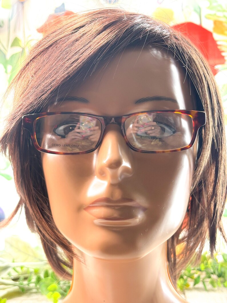 Puede incluir: Gafas rectangulares con estampado de carey. Las monturas marrones y &aacute;mbar tienen el texto "DEMO LENS ONLY" en la lente. Las gafas se muestran en una cabeza de maniqu&iacute; con cabello casta&ntilde;o.