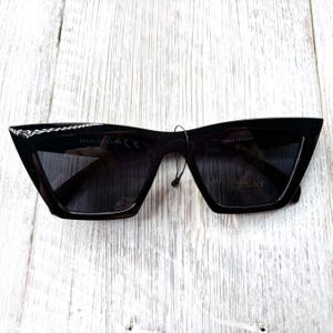 Puede incluir: Gafas de sol negras con un dise&ntilde;o de montura angular &uacute;nico. Las gafas tienen lentes oscuras y una peque&ntilde;a etiqueta adherida a la montura. La montura tiene el texto "54021 145 CAT3" y "SHINA NFASHT".