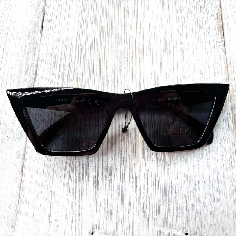 Puede incluir: Gafas de sol negras con un dise&ntilde;o de montura angular &uacute;nico. Las gafas tienen lentes oscuras y una peque&ntilde;a etiqueta adherida a la montura. La montura tiene el texto "54021 145 CAT3" y "SHINA NFASHT".