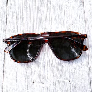 May include: Tortoiseshell sunglasses with dark lenses. The sunglasses have a classic, rectangular frame design. The arms and frame feature a brown and black pattern. The sunglasses are laying on a white wooden surface.