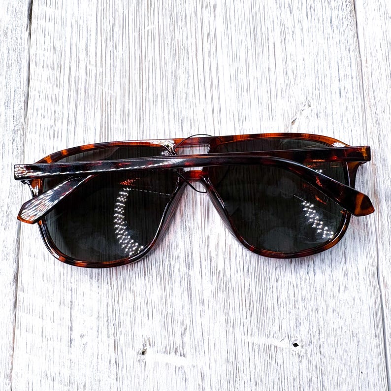 May include: Tortoiseshell sunglasses with dark lenses. The sunglasses have a classic, rectangular frame design. The arms and frame feature a brown and black pattern. The sunglasses are laying on a white wooden surface.