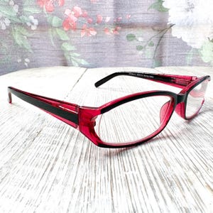 Puede incluir: Gafas rojas y negras con montura rectangular. Las gafas tienen lentes transparentes y una franja negra en las patillas. El fondo es una superficie de madera clara con un estampado floral.