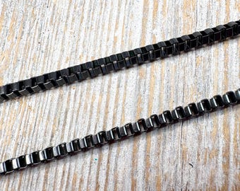 May include: Close-up of two black metal box chain necklaces. The chains are made of small, rectangular links and are displayed on a light-coloured, textured wooden surface. The chains are a dark, matte black colour.