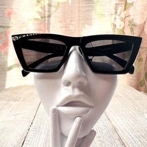 Puede incluir: Gafas de sol negras con montura geom&eacute;trica y lentes oscuras. Las gafas de sol tienen un elemento decorativo en la parte superior de la montura. Las gafas se exhiben en una cabeza de maniqu&iacute; blanca.
