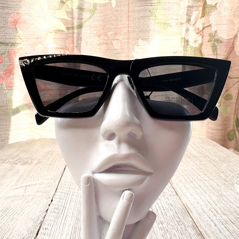 Puede incluir: Gafas de sol negras con montura geom&eacute;trica y lentes oscuras. Las gafas de sol tienen un elemento decorativo en la parte superior de la montura. Las gafas se exhiben en una cabeza de maniqu&iacute; blanca.