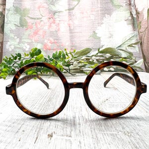 May include: Round, tortoiseshell-patterned spectacles with clear lenses. The glasses feature a classic, retro design with a keyhole bridge. The frames are a warm brown colour with darker flecks, and the temples are thin and brown.