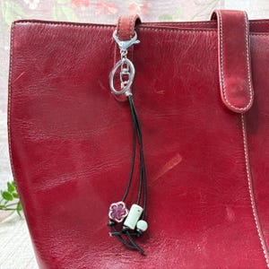 May include: A red leather tote bag with a decorative charm. The charm features a silver clasp, black cords, and small decorative beads in shades of pink, white, and green. The bag has a handle and a strap.