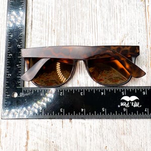 May include: Tortoiseshell sunglasses with dark lenses. The sunglasses are approximately 6 inches wide. The frame has a classic, rectangular shape. The lenses are reflective, and the sunglasses are set against a light wood background.