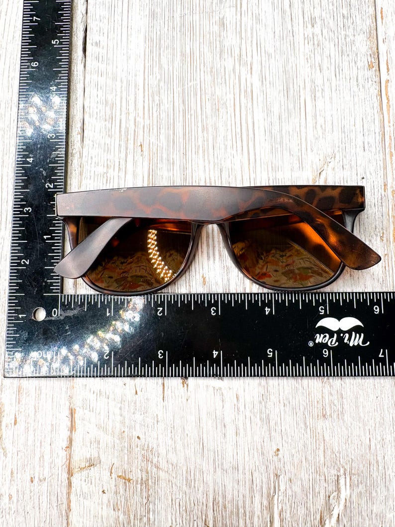 May include: Tortoiseshell sunglasses with dark lenses. The sunglasses are approximately 6 inches wide. The frame has a classic, rectangular shape. The lenses are reflective, and the sunglasses are set against a light wood background.