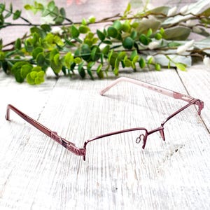 May include: A pair of rimless eyeglasses with a reddish-brown metal frame. The glasses have a rectangular shape and are resting on a white wooden surface. The temples are a lighter pink color.