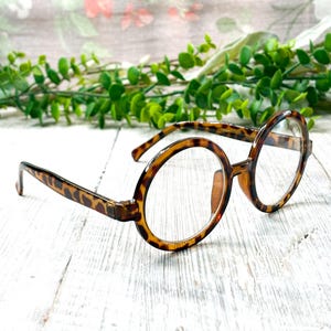 May include: Round eyeglasses with a tortoiseshell pattern. The frames are brown and tan, with clear lenses. The glasses are set against a backdrop of greenery and a white wooden surface.
