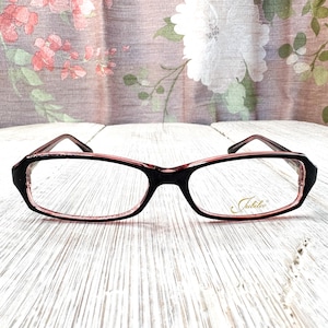 May include: Black and pink framed spectacles with a rectangular shape. The glasses have a pink interior and the brand name "Jubilee" is printed in gold on the right lens. The frames are set against a floral background.