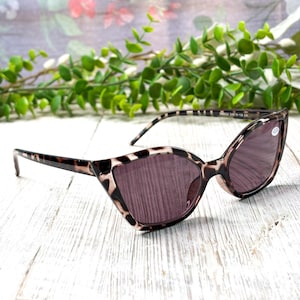 May include: Cat-eye style sunglasses with a leopard print frame and tinted lenses. The sunglasses are set against a white wooden surface with a floral background and greenery.