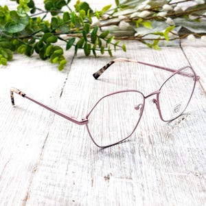 Puede incluir: Gafas con montura geométrica en tono rosa pálido. Las lentes son transparentes y las patillas presentan un diseño de carey. La marca "KED" es visible en la lente. Las gafas están sobre una superficie de madera blanca.