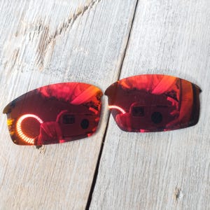 Puede incluir: Un par de lentes de repuesto para gafas de sol, de color rojo espejo. Las lentes son curvadas y tienen una superficie reflectante.