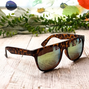 May include: Brown sunglasses with a tortoiseshell pattern on the arms and a reflective lens. The sunglasses are resting on a light-colored wooden surface, with a blurred floral background.