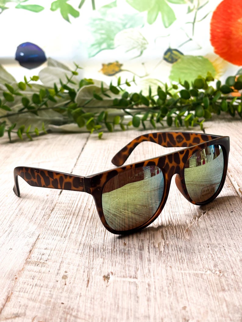 May include: Brown sunglasses with a tortoiseshell pattern on the arms and a reflective lens. The sunglasses are resting on a light-colored wooden surface, with a blurred floral background.