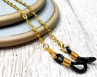 May include: Gold-colored eyeglass chain with a delicate link design. The chain connects to black eyeglass holders with gold accents. The chain is displayed on a light-colored wooden surface.