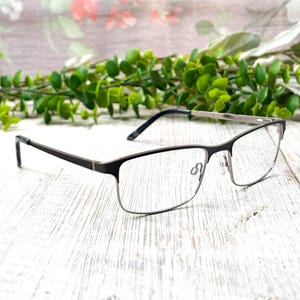 May include: Rectangular eyeglasses with a black and silver frame. The glasses have a thin, metal frame around the lenses and black temples. The background features greenery and a white wooden surface.