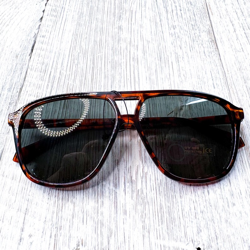 May include: Tortoiseshell aviator sunglasses with dark lenses. The sunglasses have a classic design with a double bridge and a sturdy frame. The lenses offer UV 400 protection.