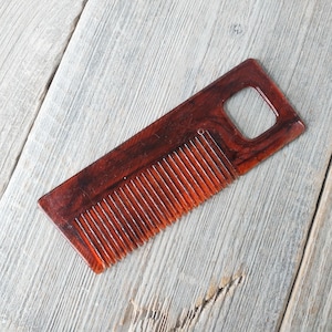 May include: A rectangular, reddish-brown wooden comb with a handle featuring a square cutout. The comb has closely spaced teeth and a smooth, polished finish. The wood grain is visible, adding to its natural aesthetic.
