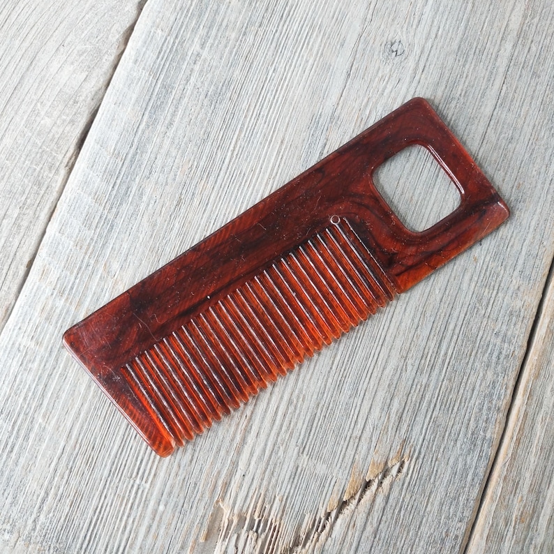 May include: A rectangular, reddish-brown wooden comb with a handle featuring a square cutout. The comb has closely spaced teeth and a smooth, polished finish. The wood grain is visible, adding to its natural aesthetic.
