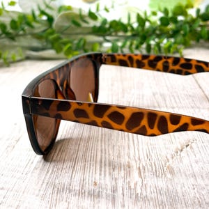 May include: Brown sunglasses with a tortoise shell pattern on the arms. The sunglasses have a classic square frame and brown lenses. The arms are a light brown with dark brown spots. The sunglasses are resting on a white wooden surface.