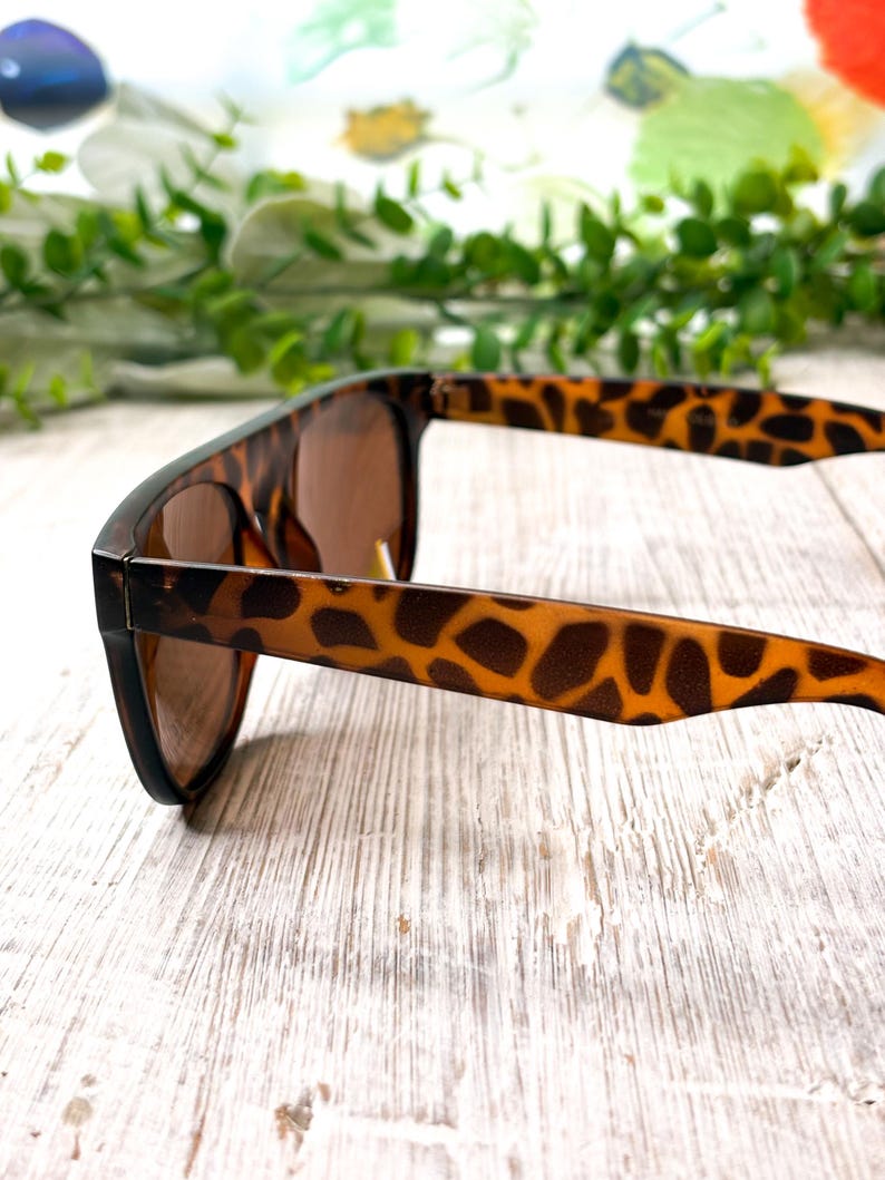 May include: Brown sunglasses with a tortoise shell pattern on the arms. The sunglasses have a classic square frame and brown lenses. The arms are a light brown with dark brown spots. The sunglasses are resting on a white wooden surface.